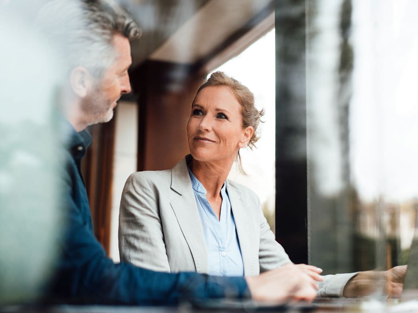 A female financial advisor in a light gray blazer and blue shirt smiling while engaged in conversation with a client in a casual blue shirt. The image is captured through a reflective surface in a professional office setting with natural lighting.