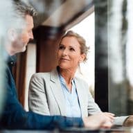 Eine Finanzberaterin in einem hellgrauen Blazer und einer blauen Bluse lächelt, während sie sich mit einem Kunden in einem lässigen blauen Hemd unterhält. Das Bild wurde durch eine reflektierende Oberfläche in einer professionellen Büroumgebung mit natürlicher Beleuchtung aufgenommen.
