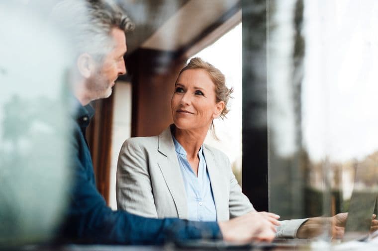 A female financial advisor in a light gray blazer and blue shirt smiling while engaged in conversation with a client in a casual blue shirt. The image is captured through a reflective surface in a professional office setting with natural lighting.