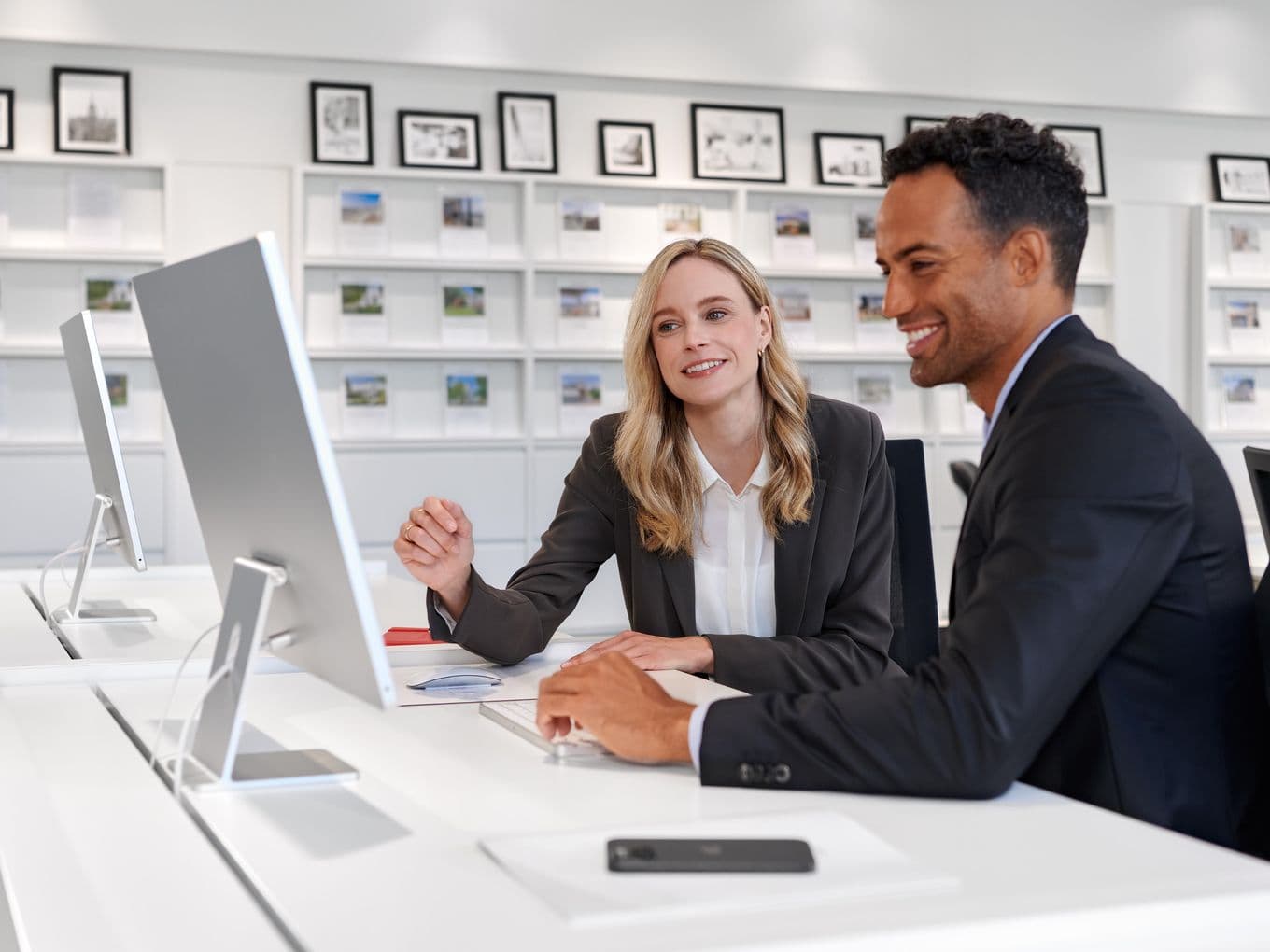 Zwei Geschäftsleute in Businesskleidung unterhalten sich an einem Computer in einem modernen, hellen Büro mit Fotos an der Wand über die Arbeit.