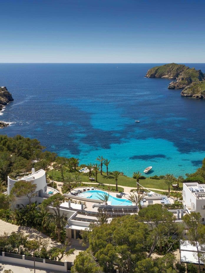 Aerial drone view of a luxury white villa with a swimming pool, nestled in a lush green forest overlooking a vibrant turquoise bay with small rocky islands under a clear blue sky.