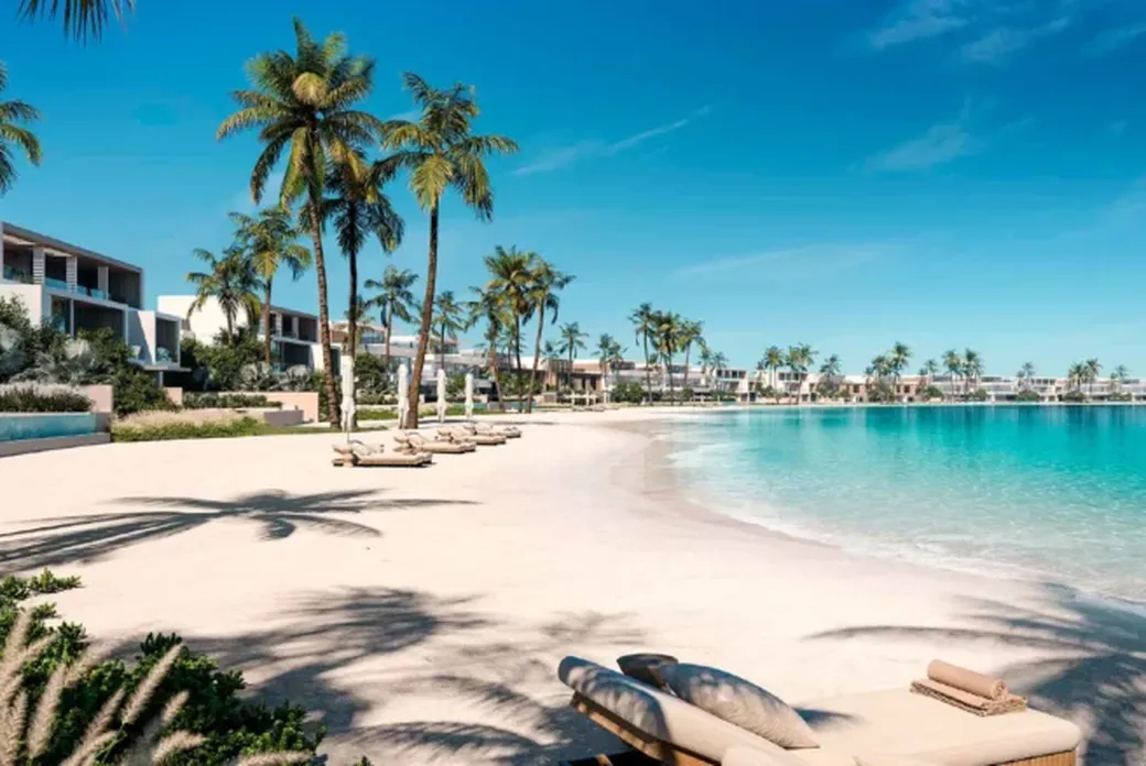 Tropical beach scene with palm trees, beachfront villas, and lounge chairs on white sand beside clear turquoise water under a clear blue sky.