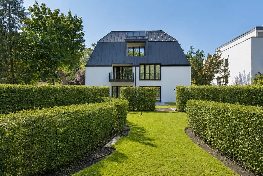 White two-story house with a dark metal roof, large windows and a balcony, framed by neatly trimmed hedges and a grassy path to the entrance. AI-generated image