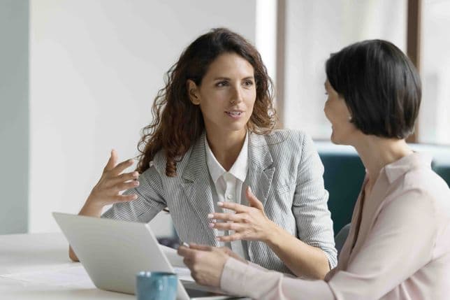Two professional women in business attire engaged in conversation at a modern office desk, one wearing a striped blazer gesturing while explaining something, the other in a light pink blazer listening attentively, with a laptop and coffee cup on the table.
