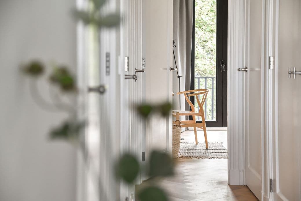 Hallway view through white doors to a light wood chair, woven basket, and black-framed window with gray curtains.