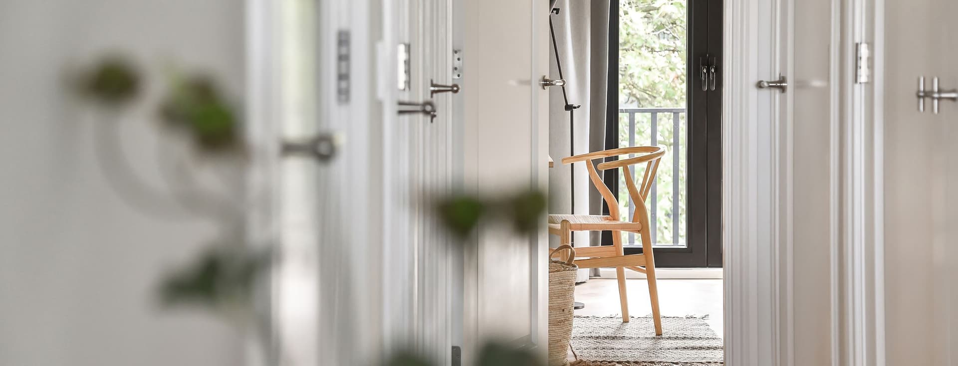 Hallway view through white doors to a light wood chair, woven basket, and black-framed window with gray curtains.