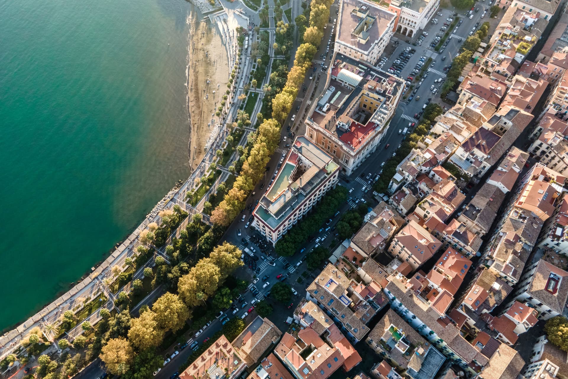 Aerial view of a coastal city with a beach, promenade, trees, buildings, and streets.