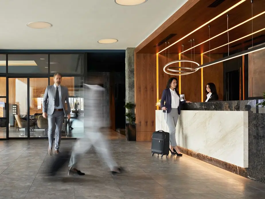 Hotel lobby with blurred figures walking, a woman with luggage at the marble reception desk, and two receptionists.