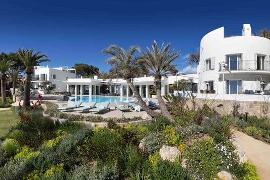 An elevated wide-angle photograph of a multi-building, Mediterranean-style white villa complex surrounding a large swimming pool and patio, set against a backdrop of a dense green pine forest and a clear blue sky.