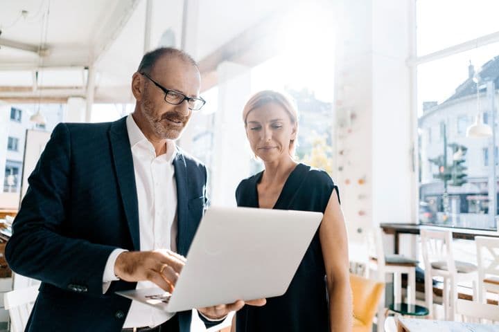 Two real estate professionals in business attire reviewing content on a laptop in a bright office space. A man wearing glasses and a dark blazer points at the screen while a woman in a black dress looks on attentively. City buildings are visible through the window in the background.