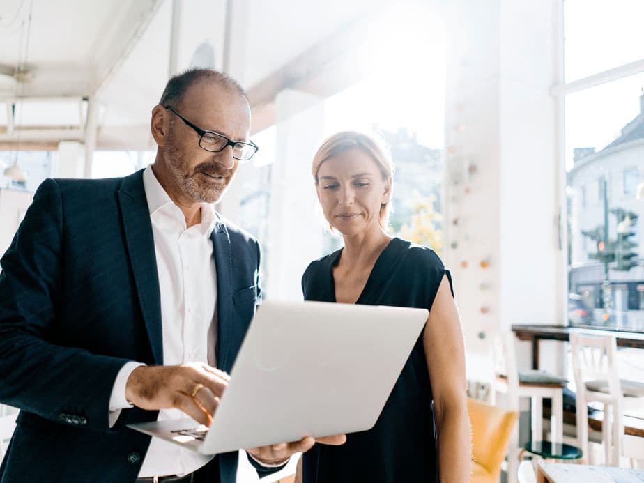 Two real estate professionals in business attire reviewing content on a laptop in a bright office space. A man wearing glasses and a dark blazer points at the screen while a woman in a black dress looks on attentively. City buildings are visible through the window in the background.