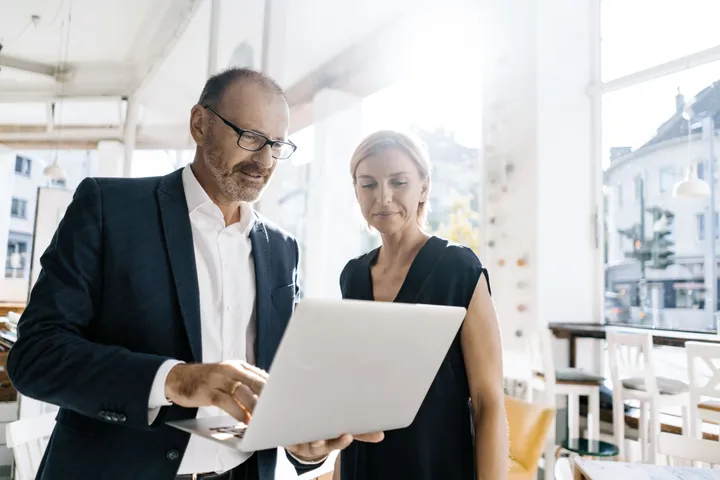 A man in a suit and a woman in black look at a laptop in a bright office.