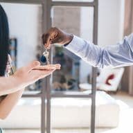 Eine lächelnde Frau erhält vor einem Fenster Schlüssel von einem Mann in blauem Hemd und Krawatte.