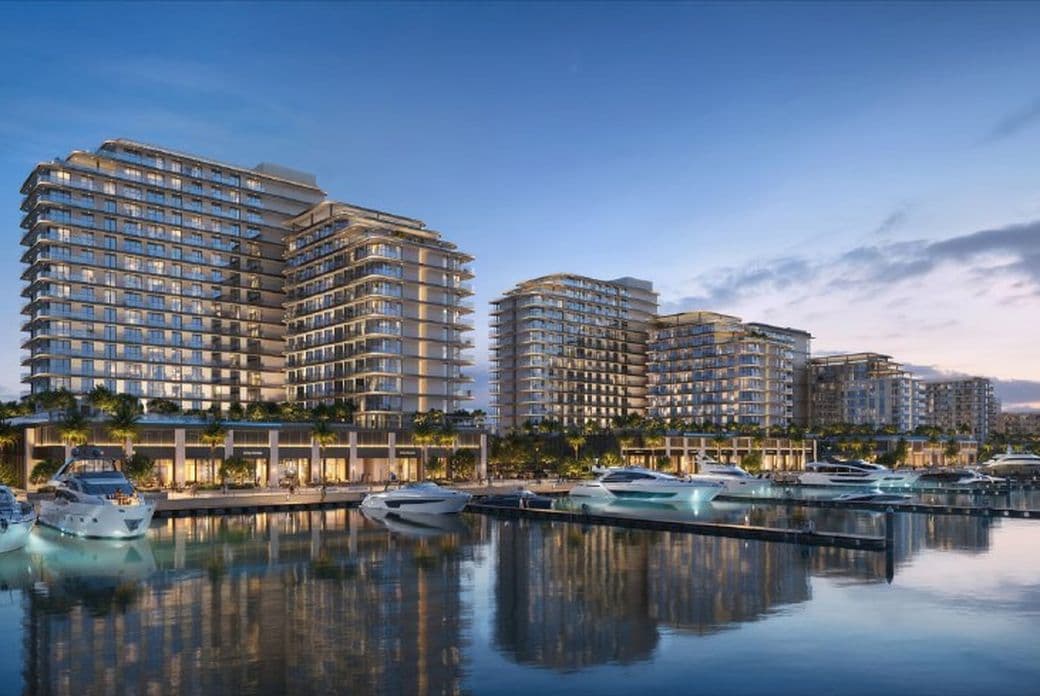Modern waterfront buildings with illuminated windows at dusk, yachts docked along the calm water reflecting the evening sky.
