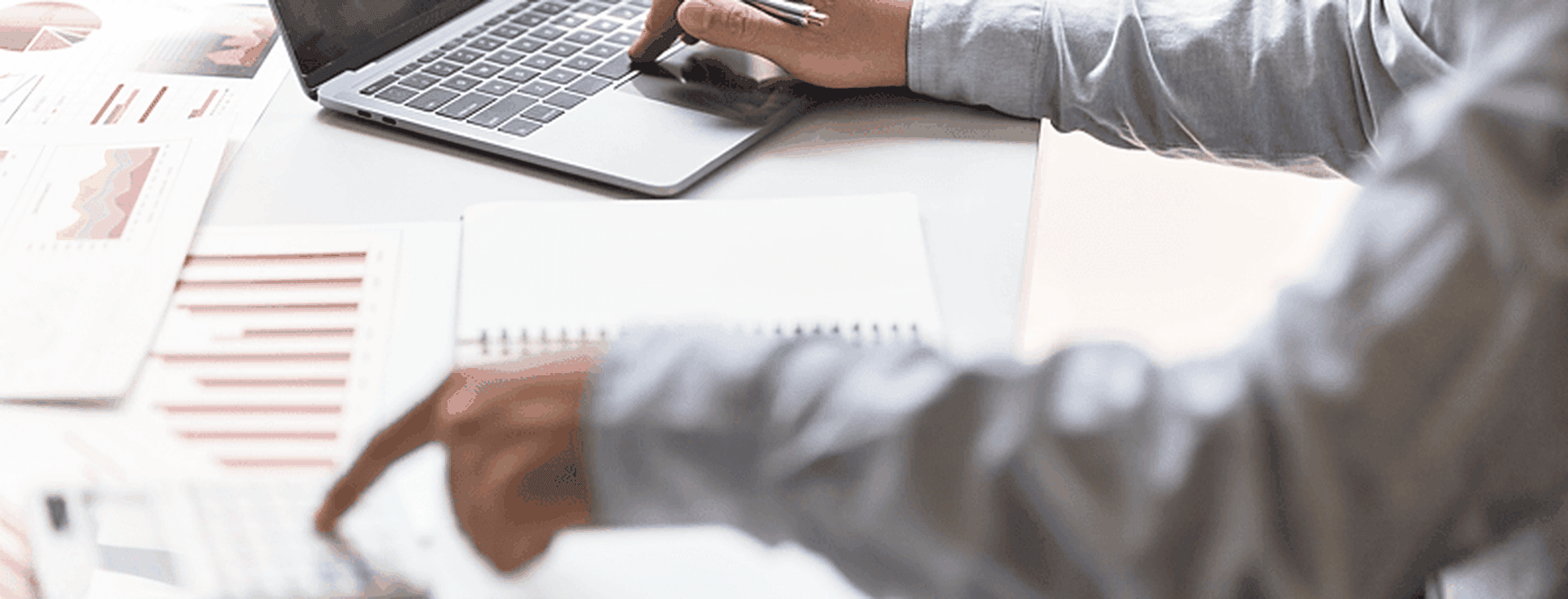 A person in a blue shirt works at a white desk with a laptop, calculator, notebook, pen, and charts.
