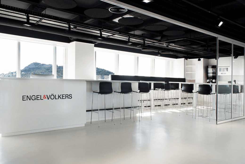 Das Büro von Engel & Völkers verfügt über ein Interieur mit einem weißen Tresen, schwarzen Hockern und großen Fenstern mit Bergblick.