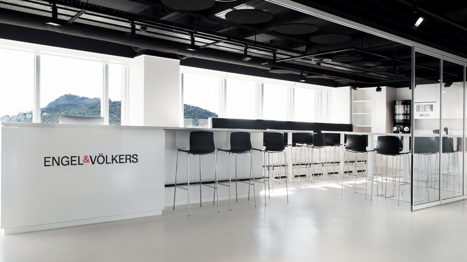Γραφείο Engel & Völkers με εσωτερικό που περιλαμβάνει λευκό πάγκο, μαύρα σκαμπό και μεγάλα παράθυρα με θέα στο βουνό.