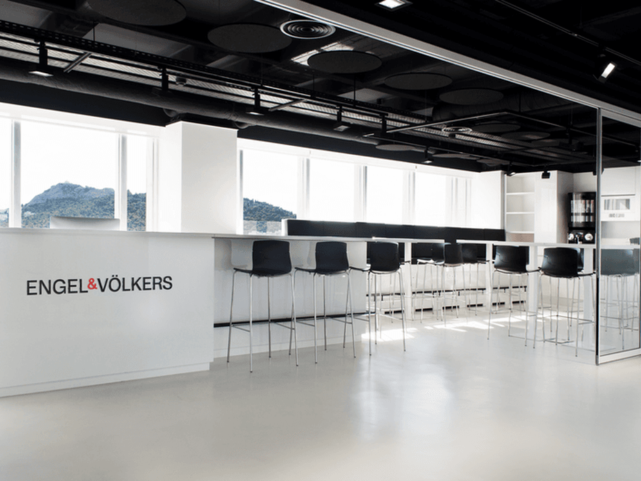 Γραφείο Engel & Völkers με εσωτερικό που περιλαμβάνει λευκό πάγκο, μαύρα σκαμπό και μεγάλα παράθυρα με θέα στο βουνό.
