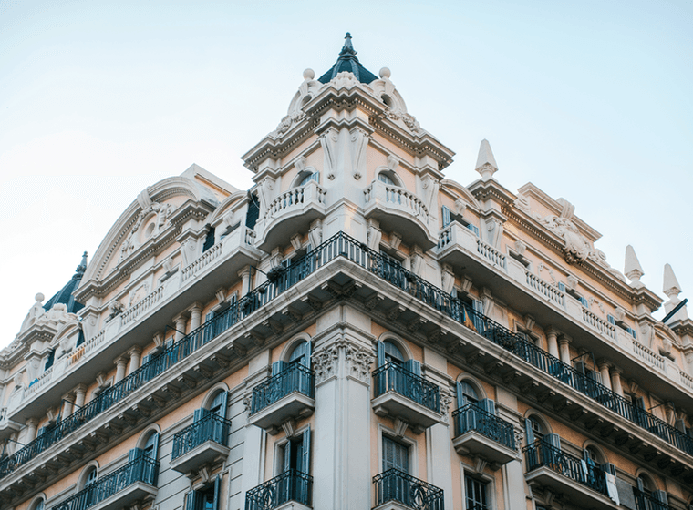 Exterior view of a tall, ornate building with balconies and blue shutters against a light blue sky.