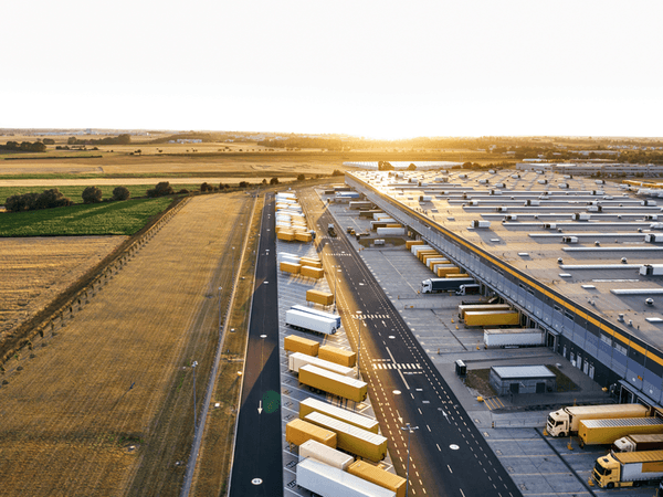 Aerial view of a large logistics center with numerous trucks and trailers in orderly loading areas, adjacent to wide fields in low sun.