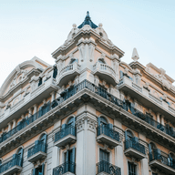Ornate multi-story building with decorative balconies, intricate architectural details, and a pointed roof against a clear blue sky.