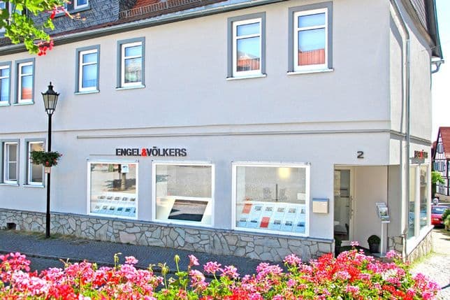 Ein weißes Gebäude mit „Engel & Völkers“-Schriftzug, großen Schaufenstern mit Angeboten und rosa Blumen im Vordergrund.