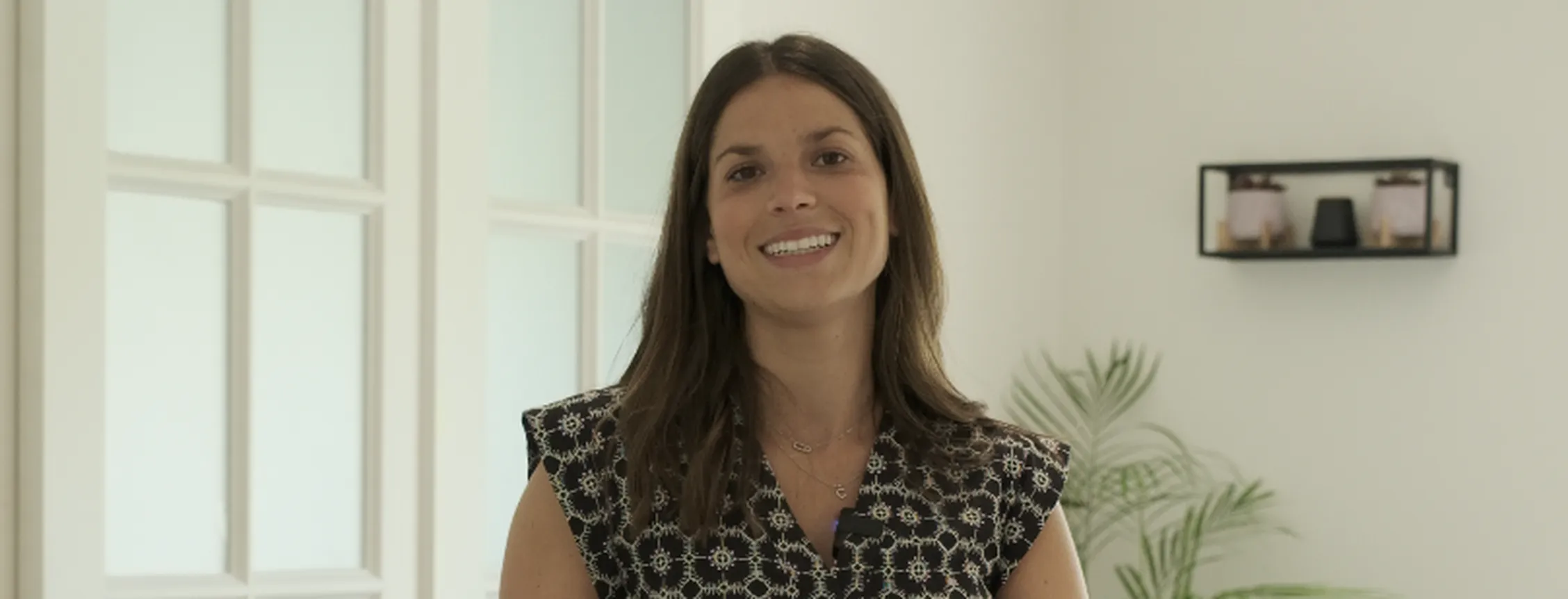 Smiling woman with shoulder-length brown hair stands indoors in a bright room, wearing a patterned sleeveless blouse, near glass doors and a plant.
