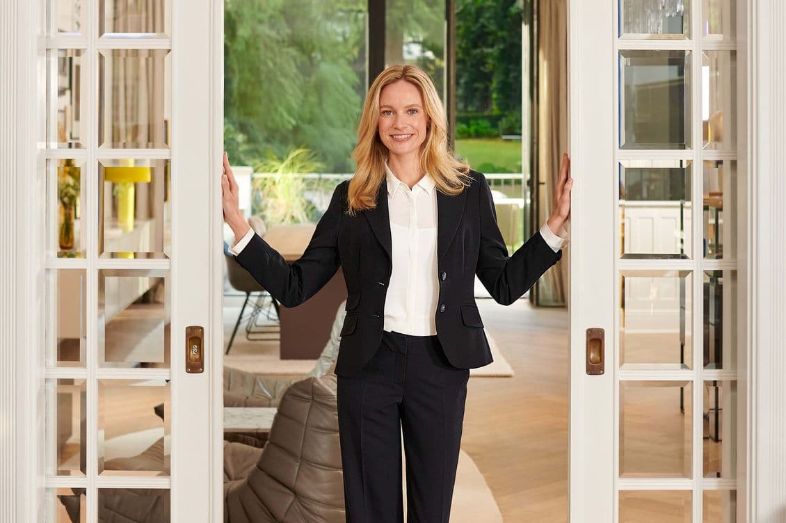 A woman in a black suit and white blouse stands smiling in a doorway, with a modern living room and garden visible in the background.