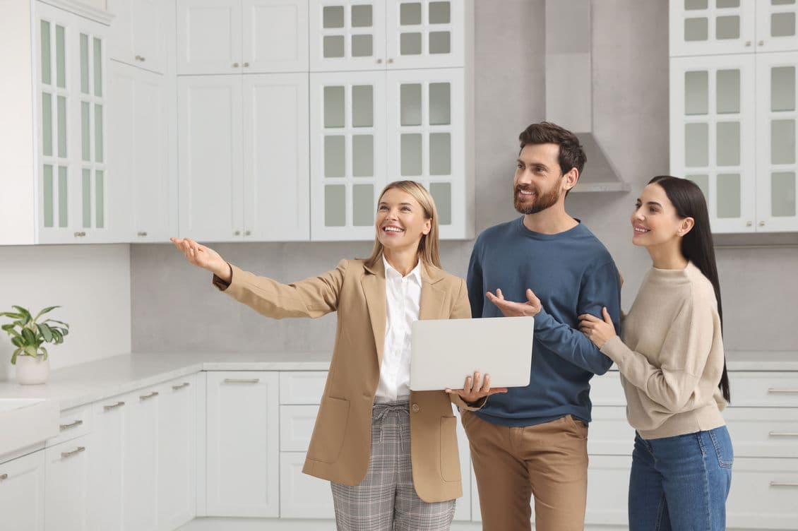 Real estate agent showing a modern kitchen to a smiling couple, with white cabinets and a potted plant on the counter.