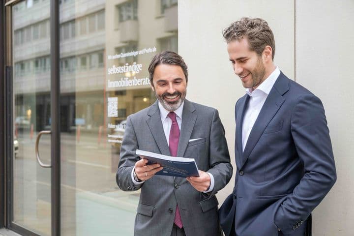 Two men in suits stand outside a building, smiling and looking at a document. A glass door and street are visible in the background.