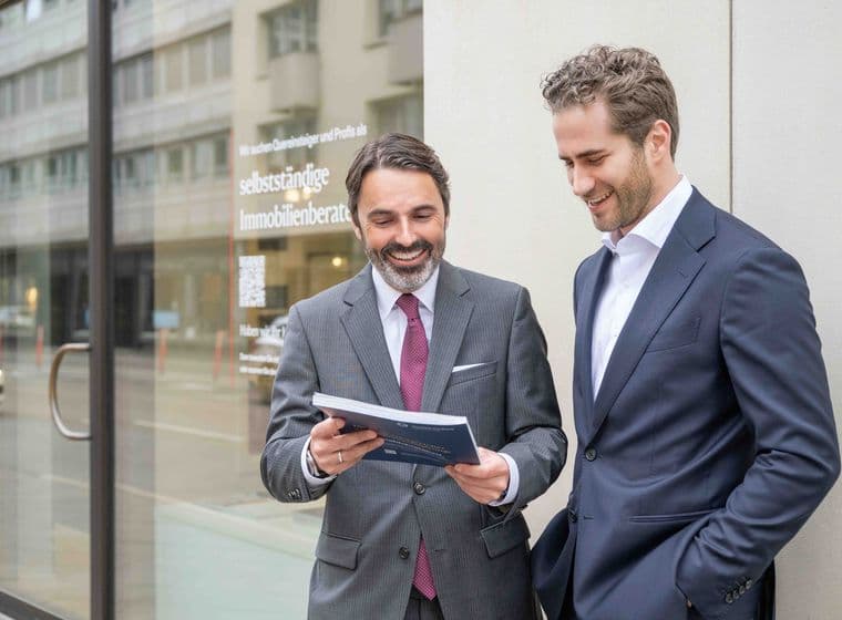 Two men in suits stand outside a building, smiling and looking at a document. A glass door and street are visible in the background.