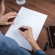 Person signing a business contract document on a wooden desk with a leather wallet and keys beside it, photographed from above