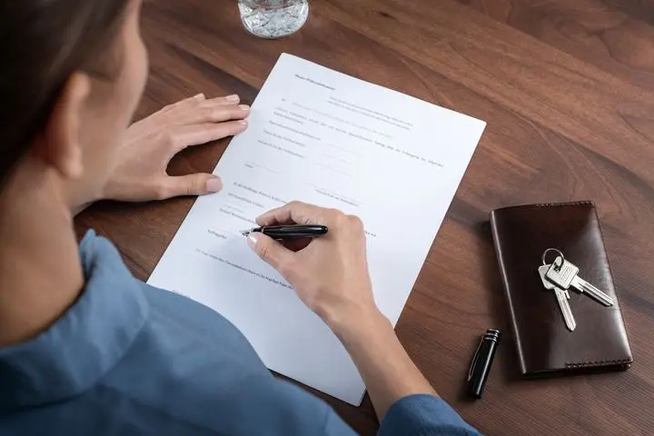 Person signing a business contract document on a wooden desk with a leather wallet and keys beside it, photographed from above