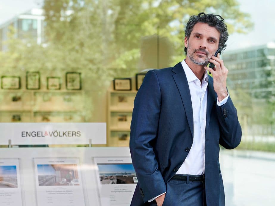 Mann im Anzug mit Krawatte, der vor einer Immobilienagentur telefoniert, mit sichtbaren Immobilienangeboten im Schaufenster.