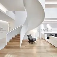 Interior view of minimalist office space featuring a white spiral staircase, wooden flooring, built-in white storage units, black rocking chair, and illustrated wall art depicting company history