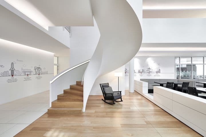 Interior view of minimalist office space featuring a white spiral staircase, wooden flooring, built-in white storage units, black rocking chair, and illustrated wall art depicting company history
