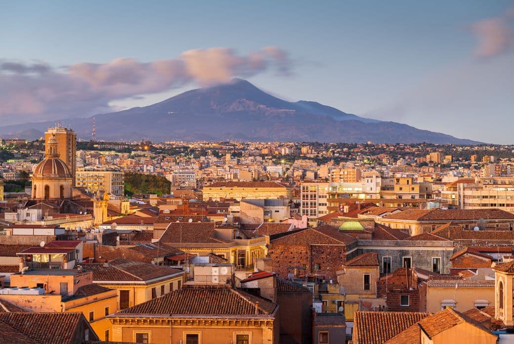Panoramic view of a city with terracotta rooftops at sunset, and a large mountain in the background partially shrouded by clouds.