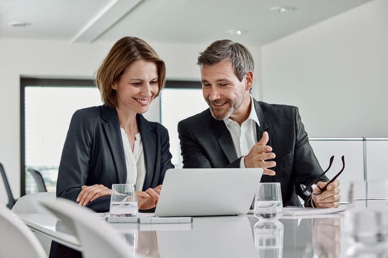 A man and woman in business attire smile while discussing something on a laptop in a bright office setting.