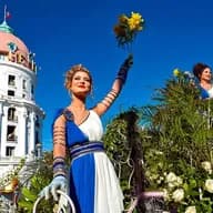A woman in a blue and white dress holds yellow flowers at the Nice Flower Parade, with a building in the background.