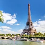 Eiffel Tower view from the Seine River, Paris. Boats docked along the riverbank, bridge in the background, blue sky with clouds.