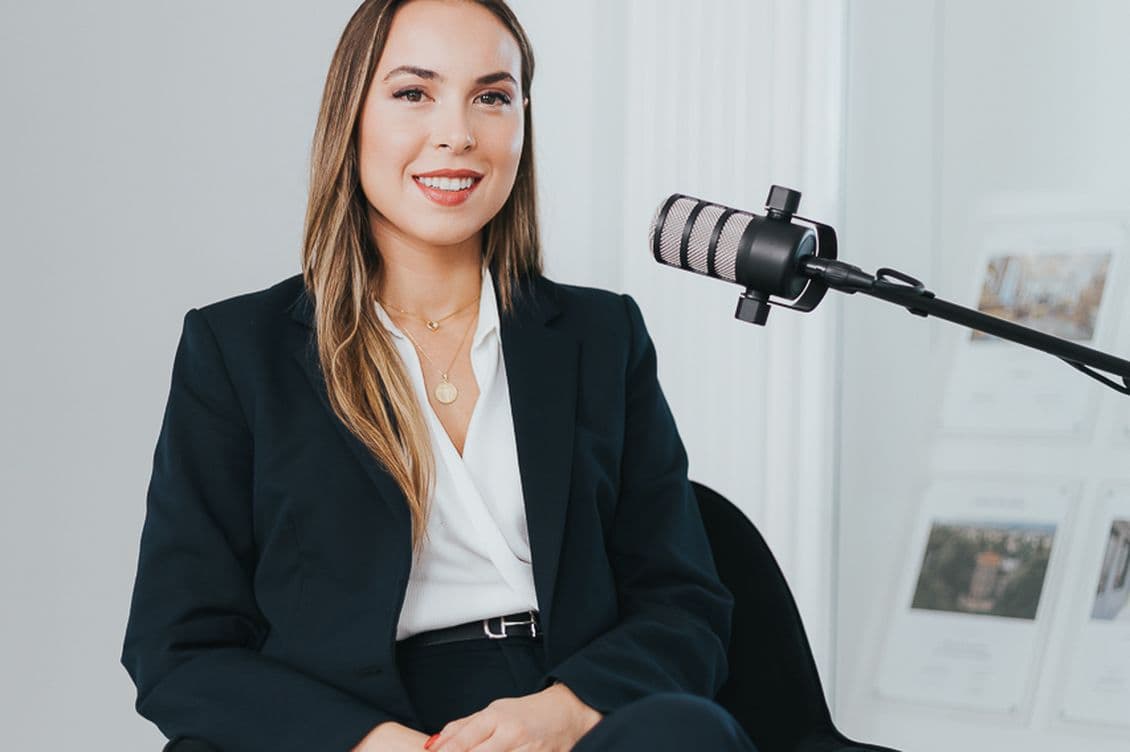 A woman in a navy suit sits in a black chair, smiling, with a microphone nearby, in a modern, bright room.