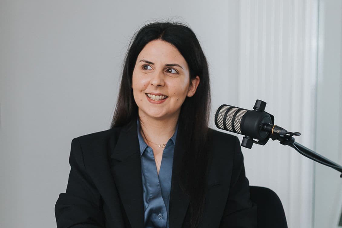 Smiling woman with long dark hair wearing a black blazer and blue shirt, sits next to a microphone, likely engaged in a conversation.