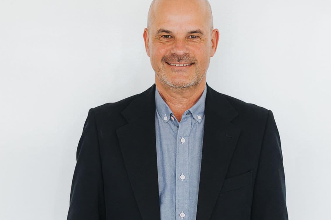 Smiling man with a bald head wearing a black blazer and blue shirt, standing against a plain white background.