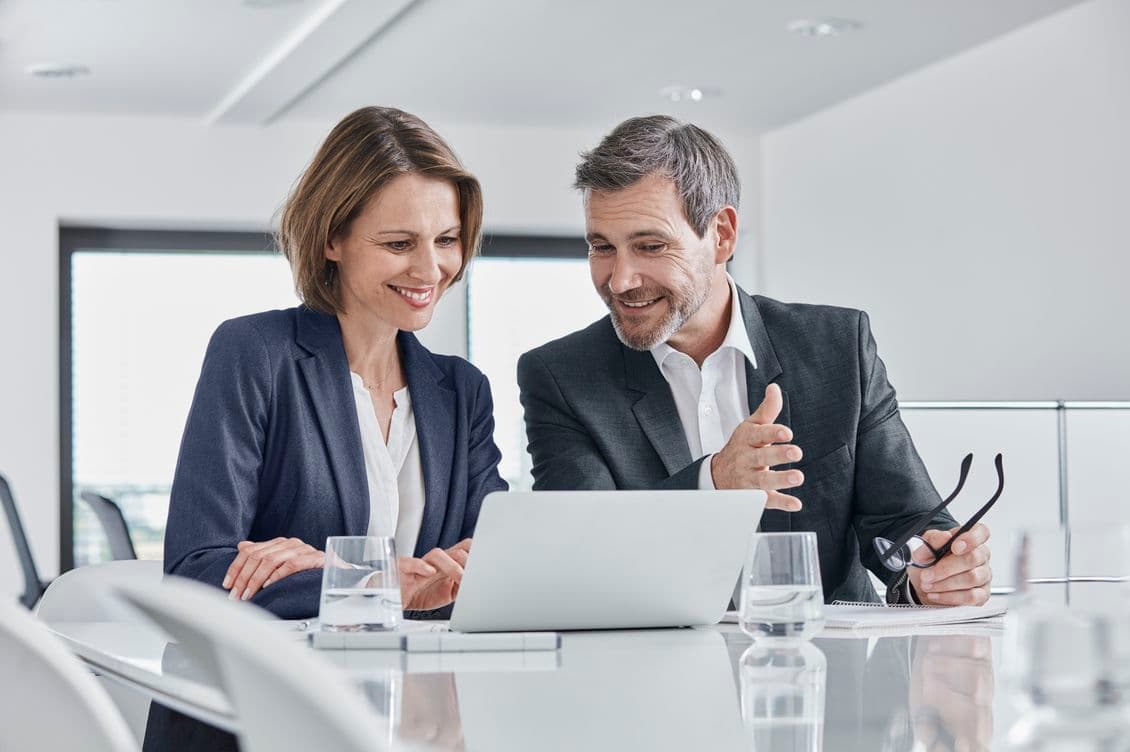 Two professionals in business attire discussing and smiling while looking at a laptop in a modern office setting.