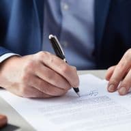 Close-up view of hands in business attire signing a formal document with a pen, showing detailed view of the signature process on white paper