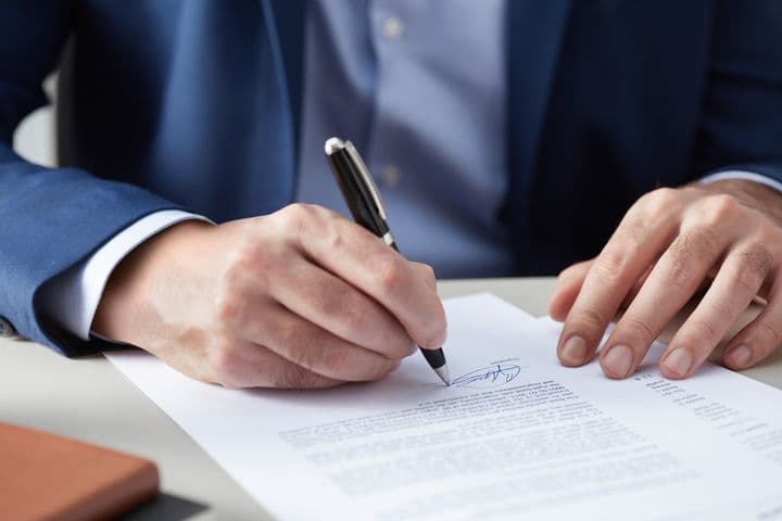 Close-up view of hands in business attire signing a formal document with a pen, showing detailed view of the signature process on white paper