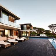 Moderne Luxusvilla mit raumhohen Glasfenstern, Infinity-Pool, Holzterrasse, Lounge-Möbeln im Freien und Panoramablick auf das Meer, umgeben von altem Baumbestand und einem wolkenverhangenen Himmel.