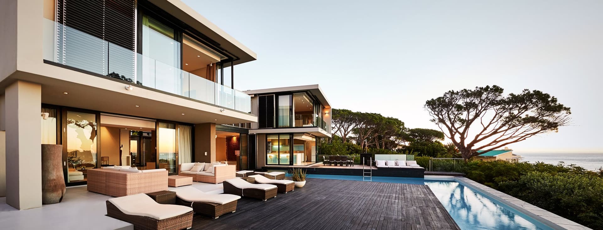 Casa moderna frente al mar con grandes ventanas, una terraza de madera, tumbonas y una piscina infinita con vista al océano al atardecer.