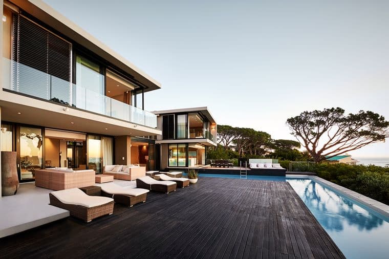 Contemporary luxury villa with floor-to-ceiling glass windows, infinity swimming pool, wooden deck terrace, outdoor lounge furniture, and panoramic ocean views with mature trees and cloudy sky backdrop
