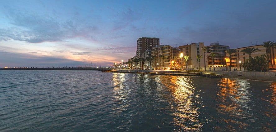Vista del port de Torrevella i la marina de nit. El mar està a l'esquerra i es pot veure un carrer il·luminat amb edificis i la posta de sol.